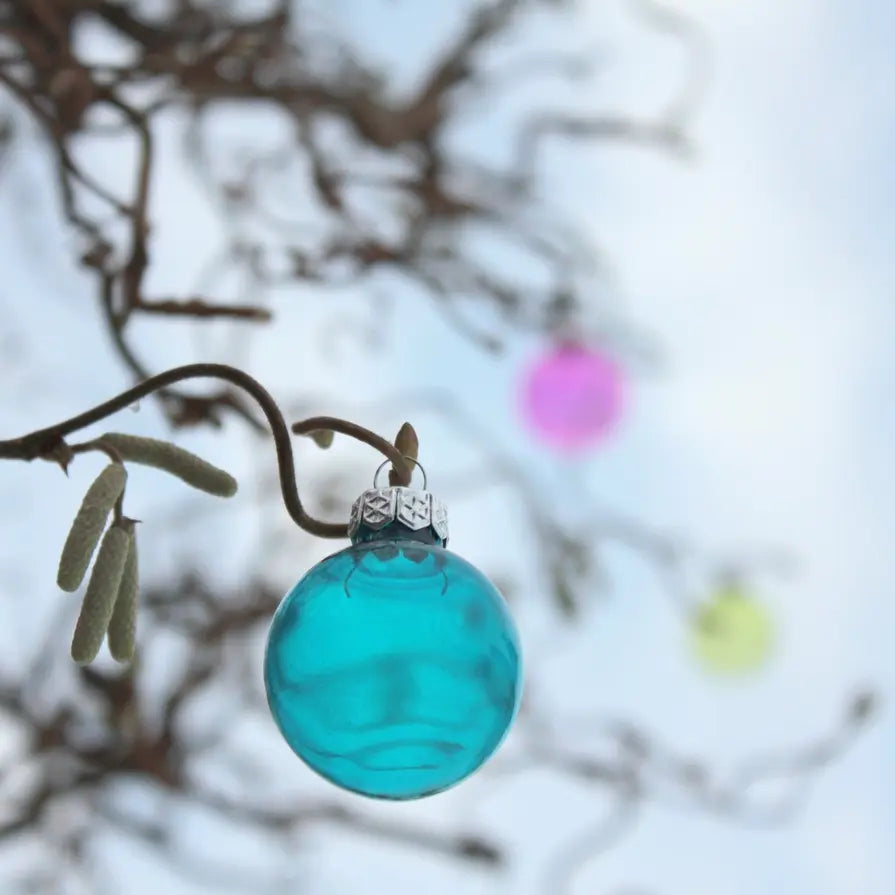 Eine blaue Weihnachtskugel hängt in einem kahlen Baum, dahinter sind Äste und der Himmel. Unscharf sind noch zwei Kugeln in Pink und Gelb zusehen.