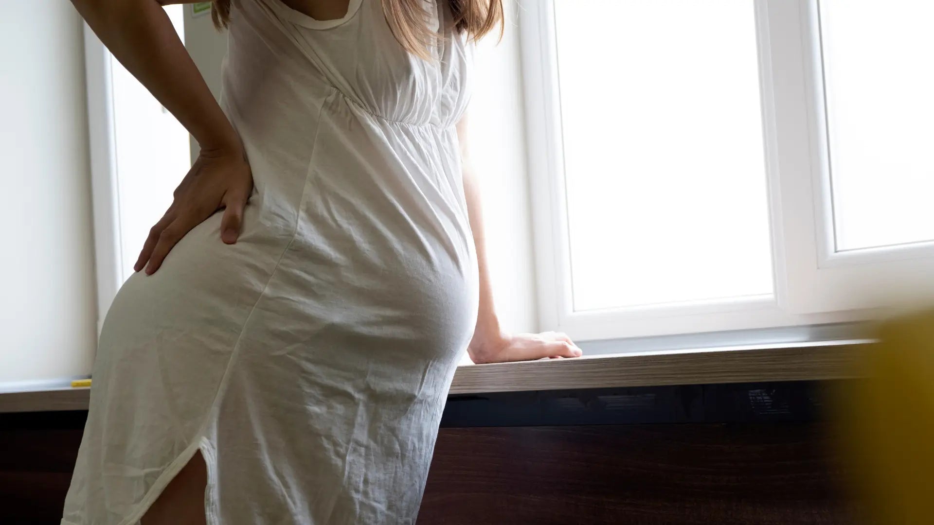 Schwangere steht am Fenster und hält sich die Hüfte